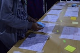 A person is arranging papers with sketches and diagrams laid out on a wooden table. Some sticky notes and markers are visible, along with various designs and wireframes. The person appears to be engaged in organizing or brainstorming.