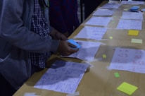 A person is arranging papers with sketches and diagrams laid out on a wooden table. Some sticky notes and markers are visible, along with various designs and wireframes. The person appears to be engaged in organizing or brainstorming.