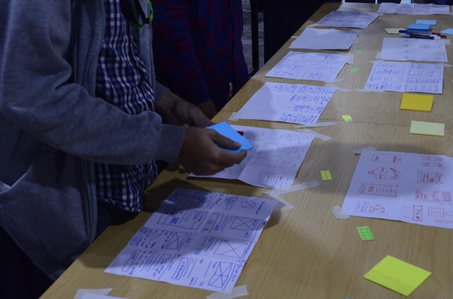 A person is arranging papers with sketches and diagrams laid out on a wooden table. Some sticky notes and markers are visible, along with various designs and wireframes. The person appears to be engaged in organizing or brainstorming.