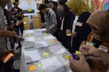 A group of people engaged in a collaborative activity around a table covered with notes, pens, and sticky notes. They appear to be working on a brainstorming or planning session. Several individuals are writing or holding sticky notes. In the background, charts and diagrams are visible on a board.