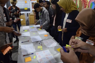 A group of people engaged in a collaborative activity around a table covered with notes, pens, and sticky notes. They appear to be working on a brainstorming or planning session. Several individuals are writing or holding sticky notes. In the background, charts and diagrams are visible on a board.