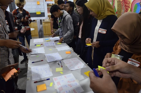 A group of people engaged in a collaborative activity around a table covered with notes, pens, and sticky notes. They appear to be working on a brainstorming or planning session. Several individuals are writing or holding sticky notes. In the background, charts and diagrams are visible on a board.