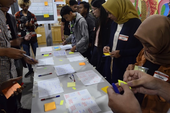 A group of people engaged in a collaborative activity around a table covered with notes, pens, and sticky notes. They appear to be working on a brainstorming or planning session. Several individuals are writing or holding sticky notes. In the background, charts and diagrams are visible on a board.