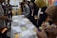 A group of people engaged in a collaborative activity around a table covered with notes, pens, and sticky notes. They appear to be working on a brainstorming or planning session. Several individuals are writing or holding sticky notes. In the background, charts and diagrams are visible on a board.