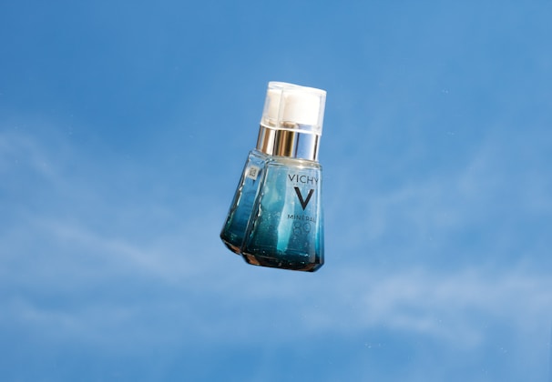 A bottle of Vichy Mineral 89 serum is photographed against a clear blue sky. The bottle is translucent with a gradient from clear to blue, and a white pump cap. The branding 'VICHY' and 'MINERAL 89' is visible on the front.