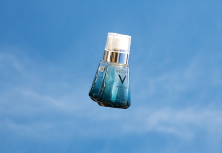 A bottle of Vichy Mineral 89 serum is photographed against a clear blue sky. The bottle is translucent with a gradient from clear to blue, and a white pump cap. The branding 'VICHY' and 'MINERAL 89' is visible on the front.