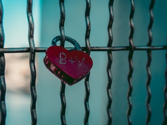 A heart-shaped red padlock with the initials B+V engraved on it is attached to a metal fence with a blurred background.