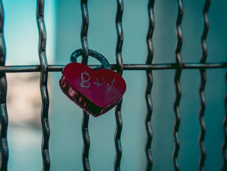 A heart-shaped red padlock with the initials B+V engraved on it is attached to a metal fence with a blurred background.