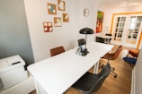 A modern office space featuring a long white table with several black and brown chairs around it. A black desk lamp and some office supplies are on the table. The wall displays abstract artwork and a clock, while a printer is located in the corner. The room has wooden flooring and a door with glass panels.