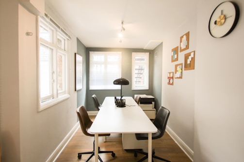 A narrow office space with a white desk at the center surrounded by two chairs in brown and black. A desk lamp is placed on the table along with some documents. The room is well-lit, featuring two windows with blinds, various wall art pieces, a black and gold wall clock, and a printer on a small cabinet.