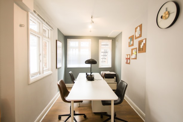 A narrow office space with a white desk at the center surrounded by two chairs in brown and black. A desk lamp is placed on the table along with some documents. The room is well-lit, featuring two windows with blinds, various wall art pieces, a black and gold wall clock, and a printer on a small cabinet.