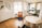 A modern, minimalist office space with wooden flooring and white walls. There is a vibrant orange chair with a cushion on a blue rug, next to a small black side table. A round white table with black chairs is positioned near a large window, allowing natural light to flood the room. The workspace area features white desks, black chairs, and a black lamp. A decorative wall with plants and interlocking mirrors adds a touch of style.