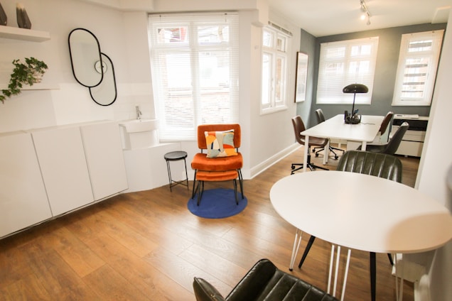 A modern, minimalist office space with wooden flooring and white walls. There is a vibrant orange chair with a cushion on a blue rug, next to a small black side table. A round white table with black chairs is positioned near a large window, allowing natural light to flood the room. The workspace area features white desks, black chairs, and a black lamp. A decorative wall with plants and interlocking mirrors adds a touch of style.