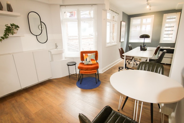 A modern, minimalist office space with wooden flooring and white walls. There is a vibrant orange chair with a cushion on a blue rug, next to a small black side table. A round white table with black chairs is positioned near a large window, allowing natural light to flood the room. The workspace area features white desks, black chairs, and a black lamp. A decorative wall with plants and interlocking mirrors adds a touch of style.