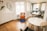 A modern, minimalist office space with wooden flooring and white walls. There is a vibrant orange chair with a cushion on a blue rug, next to a small black side table. A round white table with black chairs is positioned near a large window, allowing natural light to flood the room. The workspace area features white desks, black chairs, and a black lamp. A decorative wall with plants and interlocking mirrors adds a touch of style.