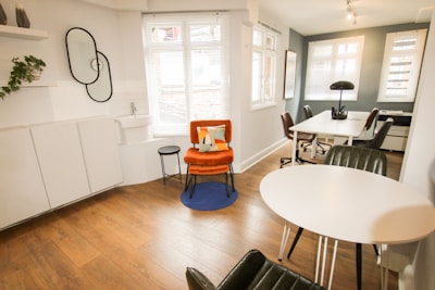 A modern, minimalist office space with wooden flooring and white walls. There is a vibrant orange chair with a cushion on a blue rug, next to a small black side table. A round white table with black chairs is positioned near a large window, allowing natural light to flood the room. The workspace area features white desks, black chairs, and a black lamp. A decorative wall with plants and interlocking mirrors adds a touch of style.