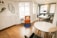 A modern, minimalist office space with wooden flooring and white walls. There is a vibrant orange chair with a cushion on a blue rug, next to a small black side table. A round white table with black chairs is positioned near a large window, allowing natural light to flood the room. The workspace area features white desks, black chairs, and a black lamp. A decorative wall with plants and interlocking mirrors adds a touch of style.