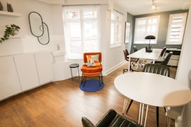 A modern, minimalist office space with wooden flooring and white walls. There is a vibrant orange chair with a cushion on a blue rug, next to a small black side table. A round white table with black chairs is positioned near a large window, allowing natural light to flood the room. The workspace area features white desks, black chairs, and a black lamp. A decorative wall with plants and interlocking mirrors adds a touch of style.