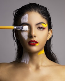 A woman with wet hair and bold red lipstick is being painted with white and yellow paint on her face and neck. A paintbrush with a yellow handle is applying white paint over her eye, adding a dynamic and artistic touch to the portrait. The background is neutral, bringing focus to her expression and the vivid colors.