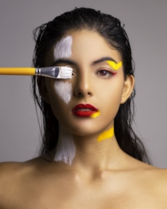 A woman with wet hair and bold red lipstick is being painted with white and yellow paint on her face and neck. A paintbrush with a yellow handle is applying white paint over her eye, adding a dynamic and artistic touch to the portrait. The background is neutral, bringing focus to her expression and the vivid colors.