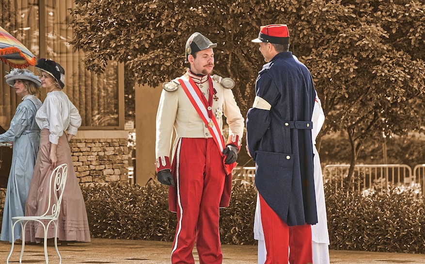 Two men dressed in historical military uniforms engage in conversation. One man wears a light-colored jacket with red accents and a matching cap, while the other man dons a dark blue coat with red and white trim and a red hat. In the background, two women in period dresses and hats are interacting near a brightly colored umbrella and a white chair. They are standing on a sidewalk with foliage and a stone wall behind them.