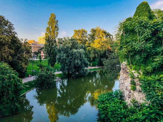 green trees beside river under blue sky during daytime in Parc des Buttes-Chaumont France