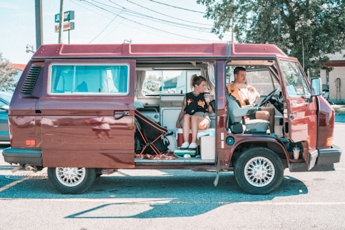 A maroon retro van is parked with its sliding door open. Inside, a woman sits on a bench seat looking at her phone, while a man sits in the driver's seat examining a map. The interior of the van shows camping equipment and supplies. The surroundings include a parking lot with cars and a 'Subway' sign in the background.