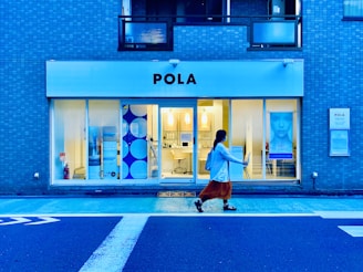 A storefront with large windows displays an interior lit with warm lighting. The exterior has a blue-tiled design, and a sign reads 'POLA' above the entrance. A person is walking past the shop on the sidewalk, wearing a white top and orange skirt.