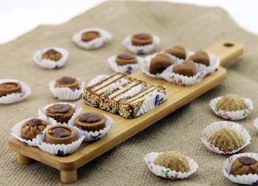 A wooden serving board on a textured surface holds an assortment of sweets, each placed in decorative paper cups. The sweets include a mix of chocolate-covered treats and nut-based confections, artfully arranged in two rows. Additional sweets are scattered around the board, creating an inviting, artisanal presentation.