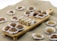 A wooden serving board on a textured surface holds an assortment of sweets, each placed in decorative paper cups. The sweets include a mix of chocolate-covered treats and nut-based confections, artfully arranged in two rows. Additional sweets are scattered around the board, creating an inviting, artisanal presentation.