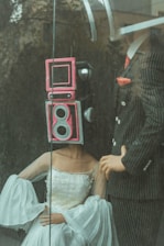 Two mannequins are posed behind a pane of glass; one is wearing a sophisticated white gown with intricate details, and has a retro camera head painted pink. The other mannequin is dressed in a pinstriped suit with a red tie, holding a confident stance.