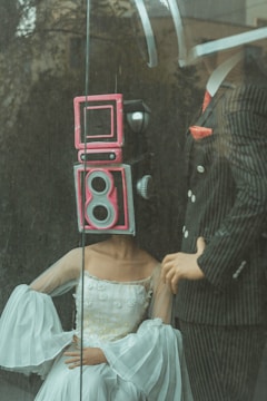 Two mannequins are posed behind a pane of glass; one is wearing a sophisticated white gown with intricate details, and has a retro camera head painted pink. The other mannequin is dressed in a pinstriped suit with a red tie, holding a confident stance.