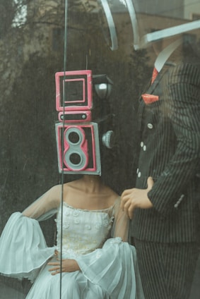 Two mannequins are posed behind a pane of glass; one is wearing a sophisticated white gown with intricate details, and has a retro camera head painted pink. The other mannequin is dressed in a pinstriped suit with a red tie, holding a confident stance.