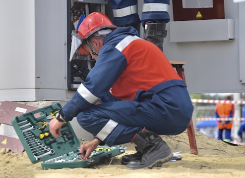 A worker in protective gear, including a red helmet and a face shield, crouches in front of an electrical panel. The worker is accessing tools from an open green toolbox on the ground. The scene takes place outdoors, with another person wearing a reflective vest standing in the background.