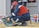 A worker in protective gear, including a red helmet and a face shield, crouches in front of an electrical panel. The worker is accessing tools from an open green toolbox on the ground. The scene takes place outdoors, with another person wearing a reflective vest standing in the background.