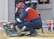 A worker in protective gear, including a red helmet and a face shield, crouches in front of an electrical panel. The worker is accessing tools from an open green toolbox on the ground. The scene takes place outdoors, with another person wearing a reflective vest standing in the background.
