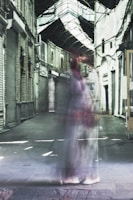 A ghostly figure walks through a narrow, covered alleyway with arched windows and brick walls, giving a surreal and haunting effect. The setting appears deserted with closed shutters.