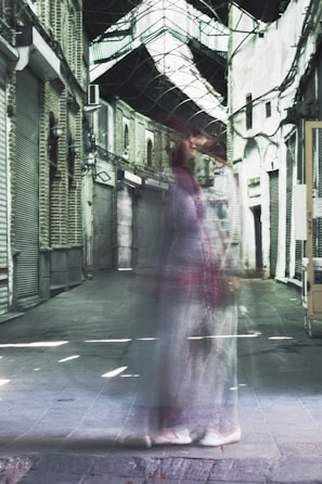 A ghostly figure walks through a narrow, covered alleyway with arched windows and brick walls, giving a surreal and haunting effect. The setting appears deserted with closed shutters.