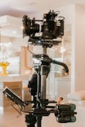 A professional video camera setup on a stabilizing rig is positioned indoors. The camera includes various components such as lenses and displays, and is mounted on a mechanical arm. The background features a home interior with soft lighting and furniture.