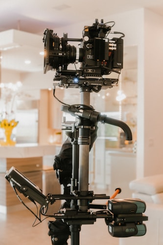 A professional video camera setup on a stabilizing rig is positioned indoors. The camera includes various components such as lenses and displays, and is mounted on a mechanical arm. The background features a home interior with soft lighting and furniture.