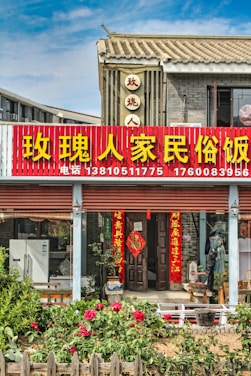 A traditional Chinese building with a red and yellow signboard displaying large Chinese characters and phone numbers. The structure features tiled roofing and grey brick walls. In front of the entrance, there are several red lanterns and door banners with Chinese calligraphy. A garden with blooming flowers and wooden fencing adorns the foreground.