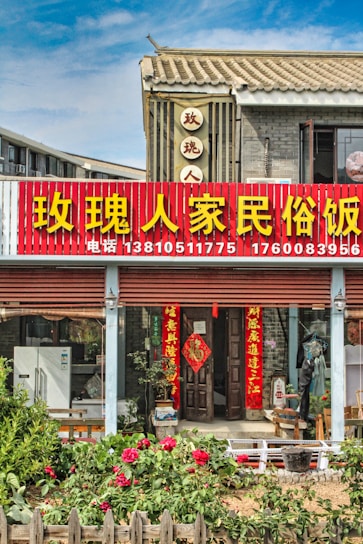 A traditional Chinese building with a red and yellow signboard displaying large Chinese characters and phone numbers. The structure features tiled roofing and grey brick walls. In front of the entrance, there are several red lanterns and door banners with Chinese calligraphy. A garden with blooming flowers and wooden fencing adorns the foreground.