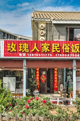 A traditional Chinese building with a red and yellow signboard displaying large Chinese characters and phone numbers. The structure features tiled roofing and grey brick walls. In front of the entrance, there are several red lanterns and door banners with Chinese calligraphy. A garden with blooming flowers and wooden fencing adorns the foreground.
