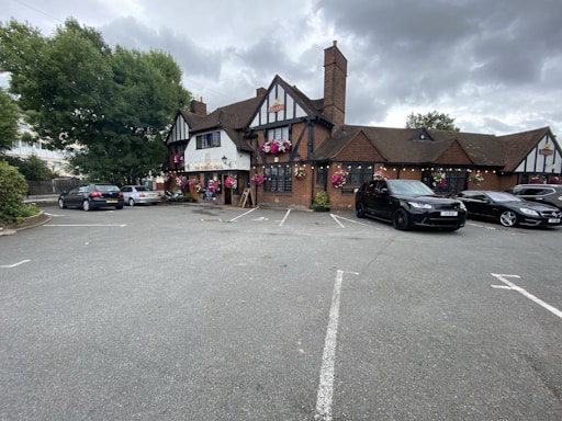 A traditional pub with a brown-tiled roof and half-timbered facade surrounded by parked cars. Hanging flower baskets add a touch of color to the building. The parking lot is mostly empty, with a few cars parked near the entrance. Large trees and overcast skies form the backdrop.