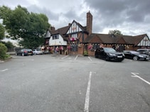 A traditional pub with a brown-tiled roof and half-timbered facade surrounded by parked cars. Hanging flower baskets add a touch of color to the building. The parking lot is mostly empty, with a few cars parked near the entrance. Large trees and overcast skies form the backdrop.