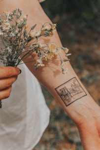 A close-up view of a person's forearm features a detailed tattoo depicting a cityscape or bridge designed like a postage stamp. The person is holding a small bouquet of dried, delicate flowers with their other hand. The background is softly blurred with earthy tones.