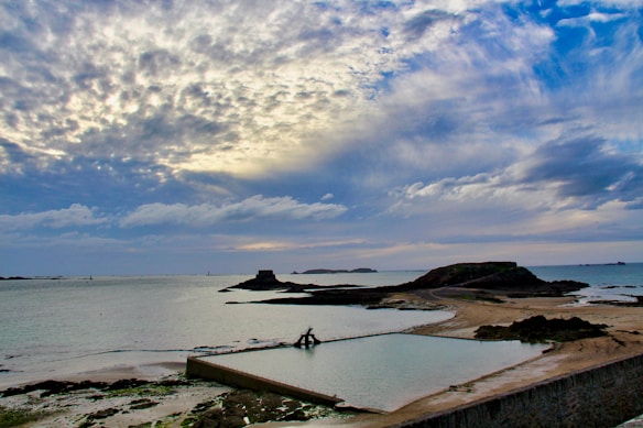 A coastal scene featuring a serene ocean with a small stone holding pool in the foreground, bordered by sandy and rocky terrain. In the distance, small rocky islands and a cloudy sky with hints of blue and gold hues create a calm and atmospheric backdrop.