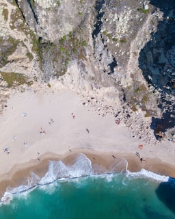 An aerial view of a serene beach bordered by rugged cliffs. Sunbathers and small groups are scattered across the sandy shore while gentle waves lap the coastline. The cliffs are rocky and interspersed with patches of greenery.