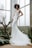 A woman stands in front of large windows wearing a sleek, white bridal gown with a fitted silhouette and a long train. The dress features delicate lace details and a plunging neckline. She is posed with one hand on her hip and the other resting on a large plant to her side. There are various types of green plants around her, including a tall, leafless tree branch and some large leafy plants. The environment appears to be an indoor garden or conservatory with natural light streaming through the windows.