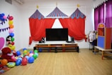 A children's play area with colorful decorations. The room features a large TV with speakers on a wooden stand. Above it is a circus-themed backdrop with red, blue, and yellow fabric resembling tents. To the left, there's a large cluster of balloons in various colors and a life-sized plush Mickey Mouse figure. The floor is strewn with more balloons and cushions. On the right, a rack displays costumes, and there's a small puppet theater booth.